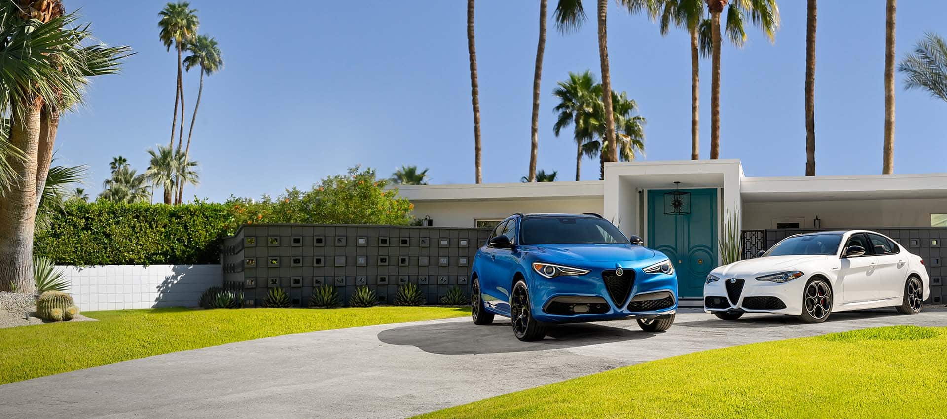A blue 2022 Alfa Romeo Stelvio TBD and white 2022 Alfa Romeo Giulia TBD parked in the driveway of a contemporary home with palm trees in the background.