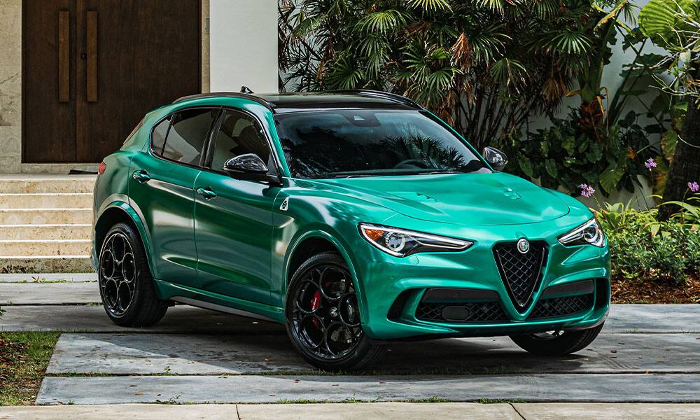 An angled front view of a green 2022 Alfa Romeo Stelvio Quadrifoglio parked in the driveway of a contemporary home with tropical greenery.