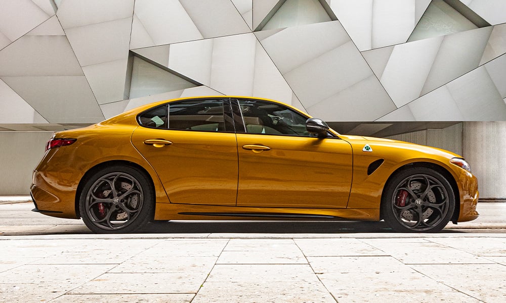 A profile view of a golden 2023 Alfa Romeo Giulia Quadrifoglio parked inside a building with an abstract design on the wall.